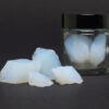 Lose Bruchstücke von Silica Aerogel mit blauem Schimmer, dahinter der Glastiegel des Produkts auf dunklem Grund